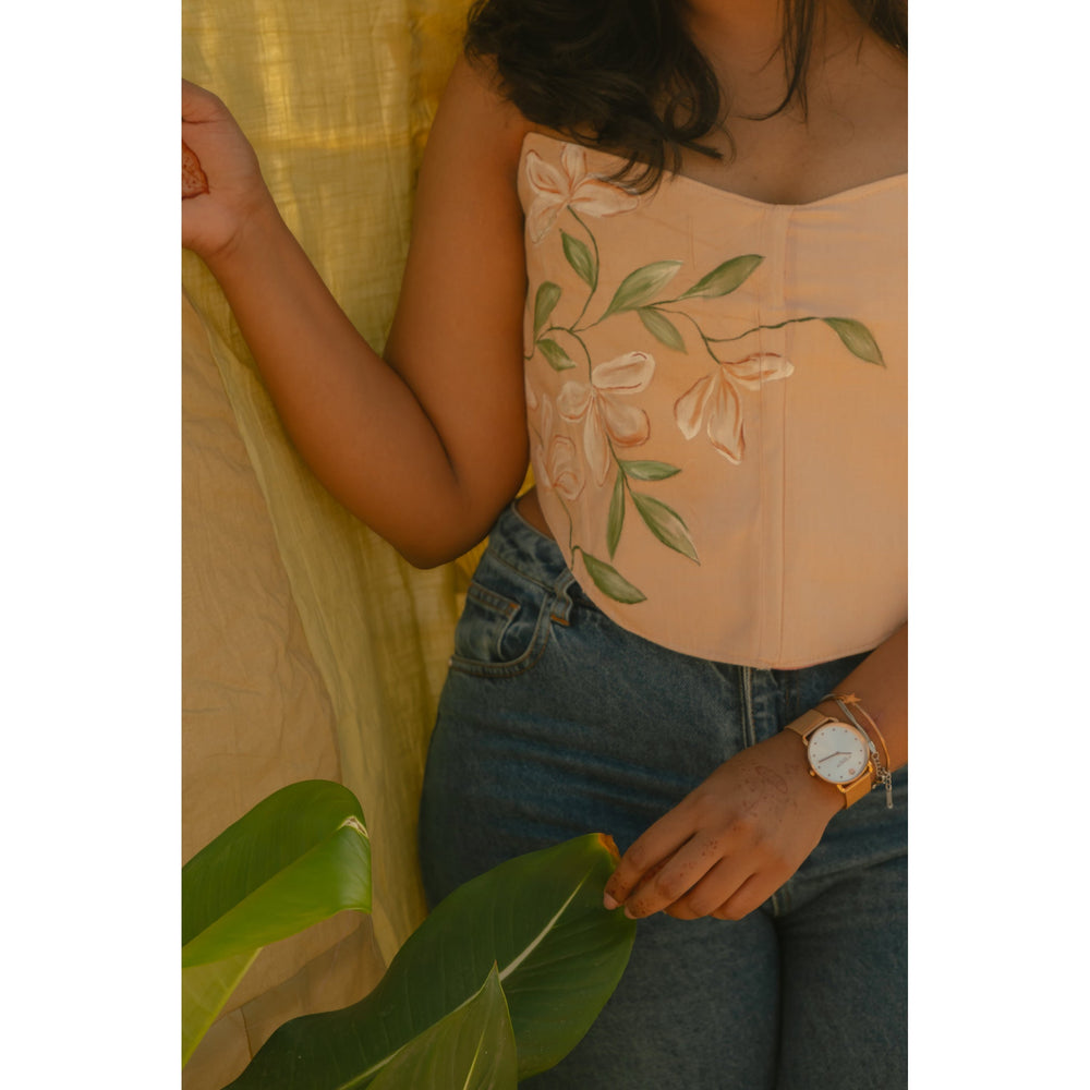 Person wearing a light pink corset top with floral hand painted details, sitting outdoors with greenery.