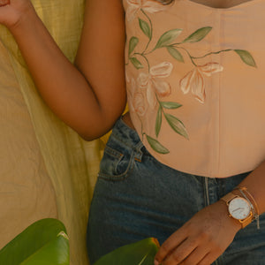 Person wearing a light pink corset top with floral hand painted details, sitting outdoors with greenery.