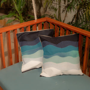 Two patterned pillows in shades of blue with a wooden railing and plants in the background